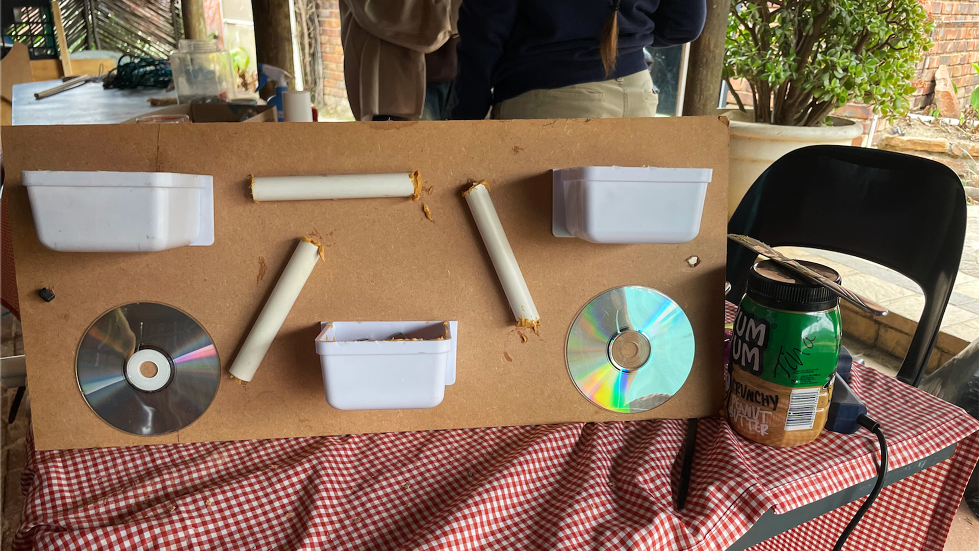A homemade contraption with containers, pipes, and CDs on a wooden board, placed on a checkered tablecloth.