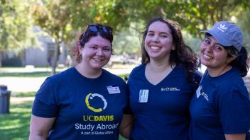 Three Global Learning Hub staff wearing blue shirts stand together in the West Quad, surrounded by greenery and enjoying the Global Learning Fair.
