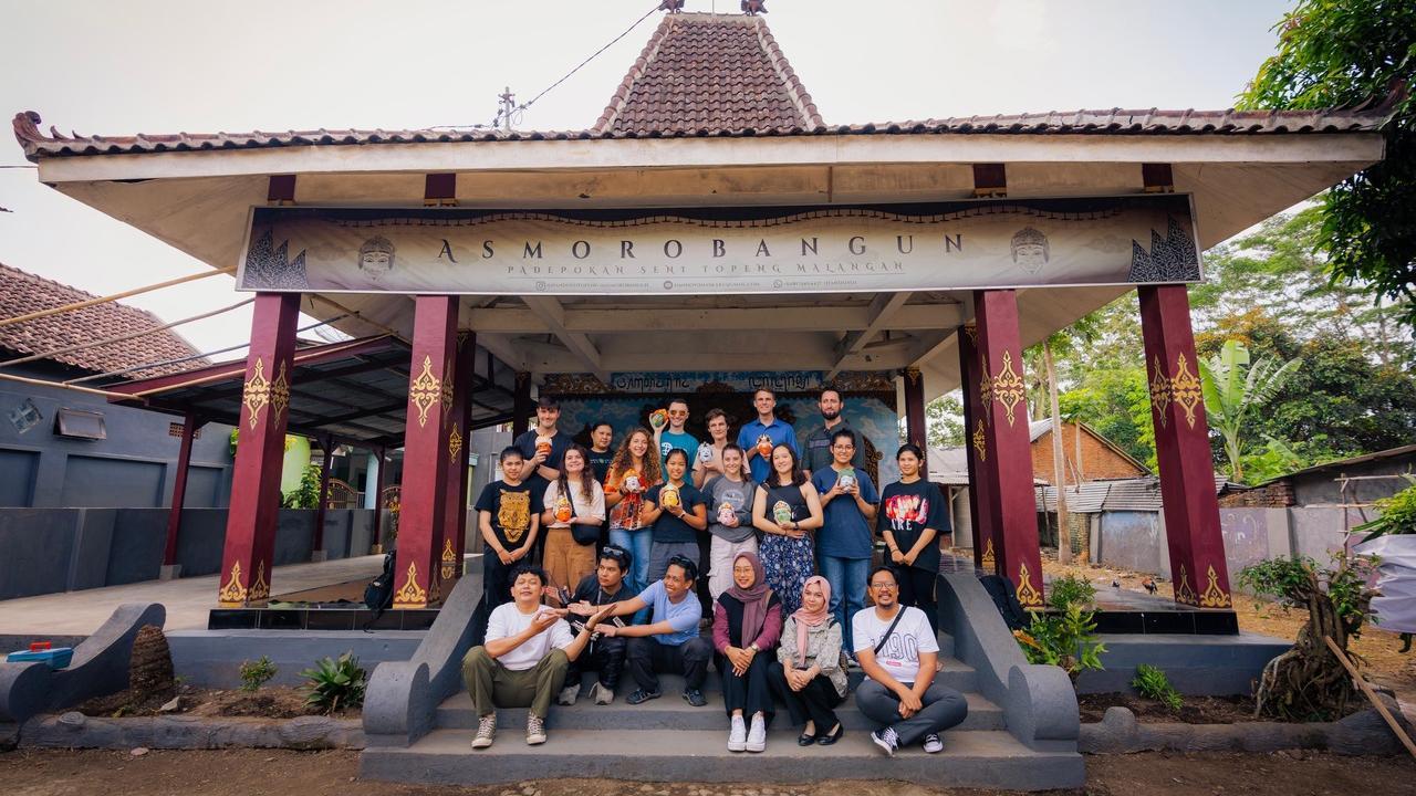 Group photo of students and locals standing and sitting together in front of the Asmorobangun cultural center in Malang, Indonesia. The building has traditional architecture with a tiled roof and decorative columns. Some individuals hold painted masks, symbolizing cultural engagement in traditional arts.