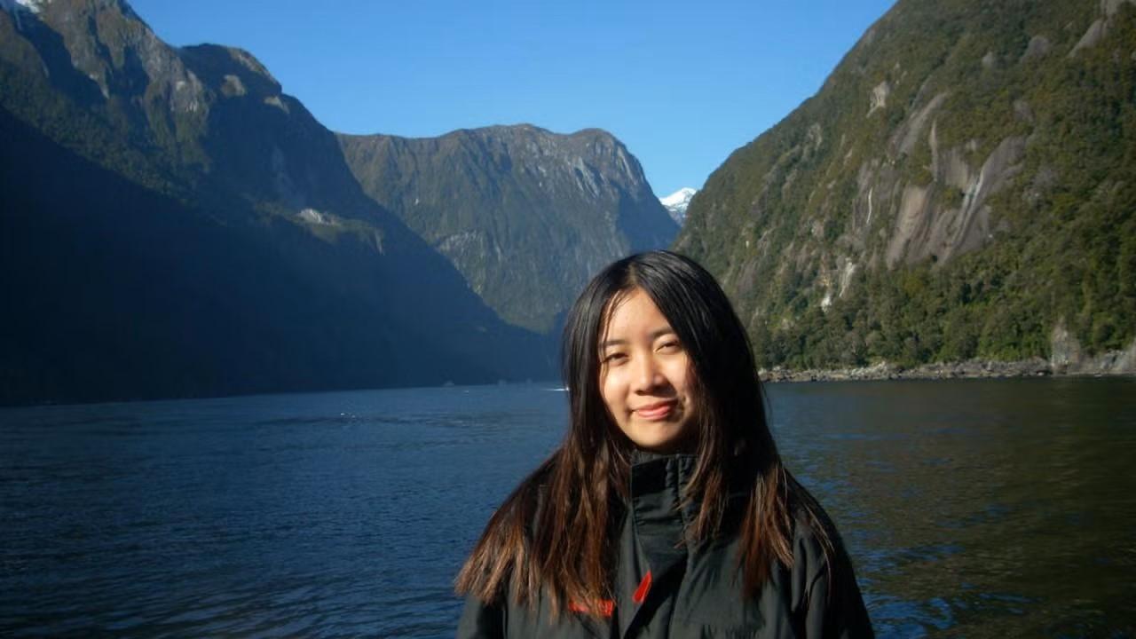 A young woman with long dark hair smiles gently while standing in front of a serene body of water surrounded by steep, forested cliffs under a clear blue sky.