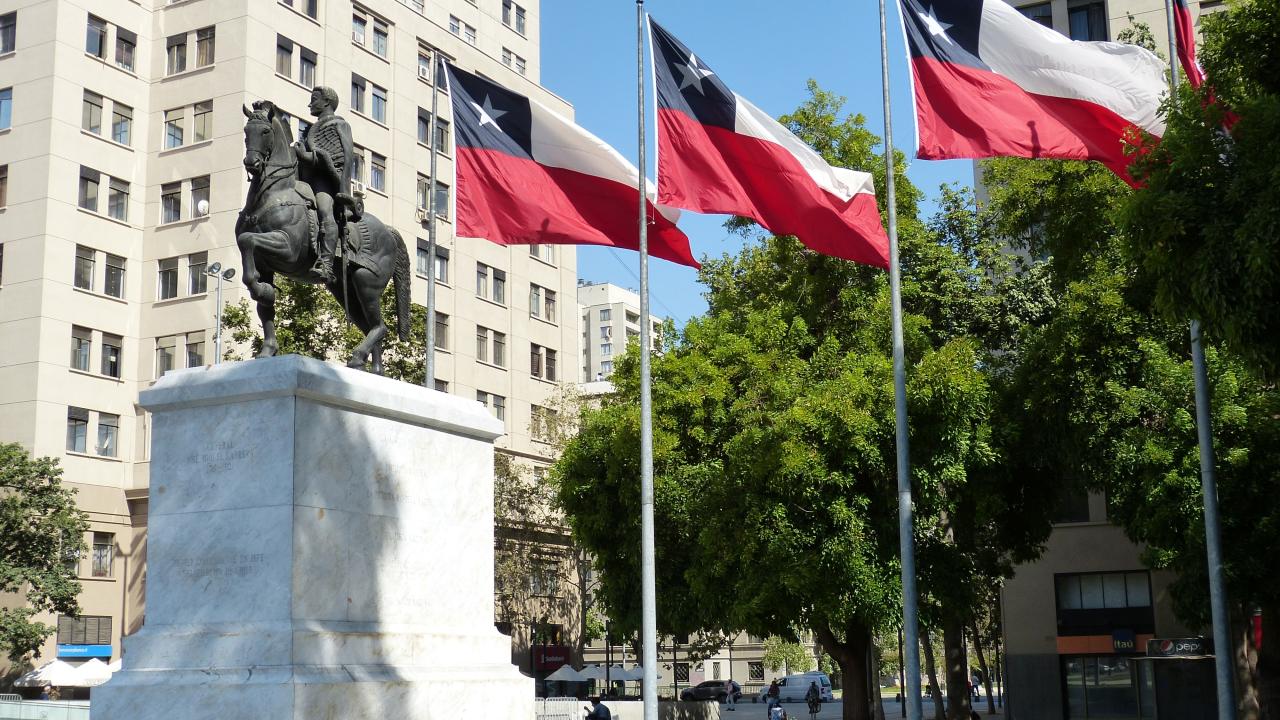 The image depicts an equestrian statue of a historical figure on a white marble pedestal. The statue is located in an urban square, surrounded by several tall, tan-colored buildings with numerous windows. To the right of the statue, four flags are displayed on tall flagpoles. Each flag features a red and white design with a blue square and a white star in the top left corner. Green trees with dense foliage are situated around the area, providing contrast to the surrounding architecture and a statuesque.