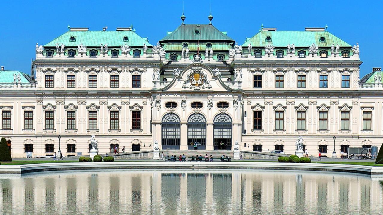 A grand baroque palace with ornate details, the Belvedere in Vienna, Austria, stands prominently with its green copper domes and symmetrical architecture. In front of the palace, a large, reflective pond mirrors the building's façade. The sky is clear and blue, and several people are visible walking along the pathways and garden areas surrounding the palace.