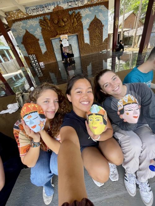 Three smiling students seated closely together, each holding a hand-painted, colorful mask. They are outdoors in front of a traditional Indonesian structure with detailed carvings, capturing a moment of cultural immersion.