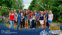 Group of student standing together and smiling into the camera. They are standing in front of a eating area and are surrounded by green trees and bushes.