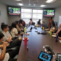 Image of students in a classroom, collaborating. Two tv screens are in the background with students participating remotely