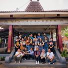 Group photo of students and locals standing and sitting together in front of the Asmorobangun cultural center in Malang, Indonesia. The building has traditional architecture with a tiled roof and decorative columns. Some individuals hold painted masks, symbolizing cultural engagement in traditional arts.