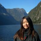 A young woman with long dark hair smiles gently while standing in front of a serene body of water surrounded by steep, forested cliffs under a clear blue sky.