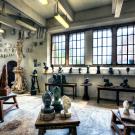 A spacious artist's studio filled with various sculptures and busts. The room has large, grid-like windows letting in natural light. Numerous small busts are arranged on a long shelf along the wall, while a variety of sculpted body parts, including arms and legs, hang on the wall as decoration. The center of the room features several stone and marble sculptures on wooden tables, including large carved figures and intricate designs.