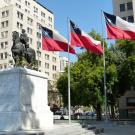 The image depicts an equestrian statue of a historical figure on a white marble pedestal. The statue is located in an urban square, surrounded by several tall, tan-colored buildings with numerous windows. To the right of the statue, four flags are displayed on tall flagpoles. Each flag features a red and white design with a blue square and a white star in the top left corner. Green trees with dense foliage are situated around the area, providing contrast to the surrounding architecture and a statuesque.