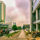 A city street scene in Kigali, Rwanda, featuring modern high-rise buildings with reflective glass facades on both sides. Motorcyclists and a few pedestrians navigate the clean, well-maintained road bordered by manicured hedges and sidewalks. The sky is overcast with gray clouds, adding a dramatic backdrop to the urban landscape. In the distance, more buildings, including a distinctively shaped skyscraper, contribute to the city's skyline.