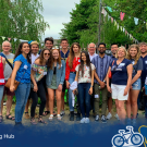 Group of student standing together and smiling into the camera. They are standing in front of a eating area and are surrounded by green trees and bushes.