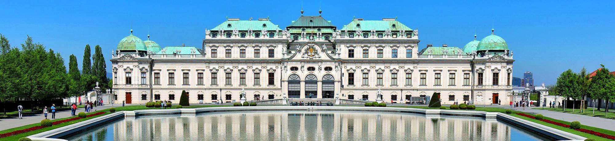 A grand baroque palace with ornate details, the Belvedere in Vienna, Austria, stands prominently with its green copper domes and symmetrical architecture. In front of the palace, a large, reflective pond mirrors the building's façade. The sky is clear and blue, and several people are visible walking along the pathways and garden areas surrounding the palace.