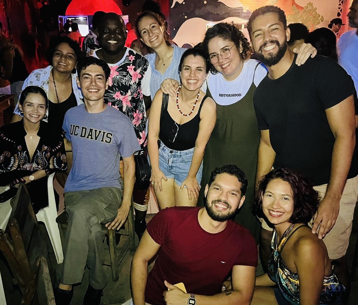 A group of ten people posing together indoors, smiling at the camera. The group consists of a diverse mix of men and women, with one person wearing a UC Davis t-shirt seated in the front row. The setting appears casual and lively, with dim lighting and colorful murals on the walls in the background, suggesting a social gathering or celebration.