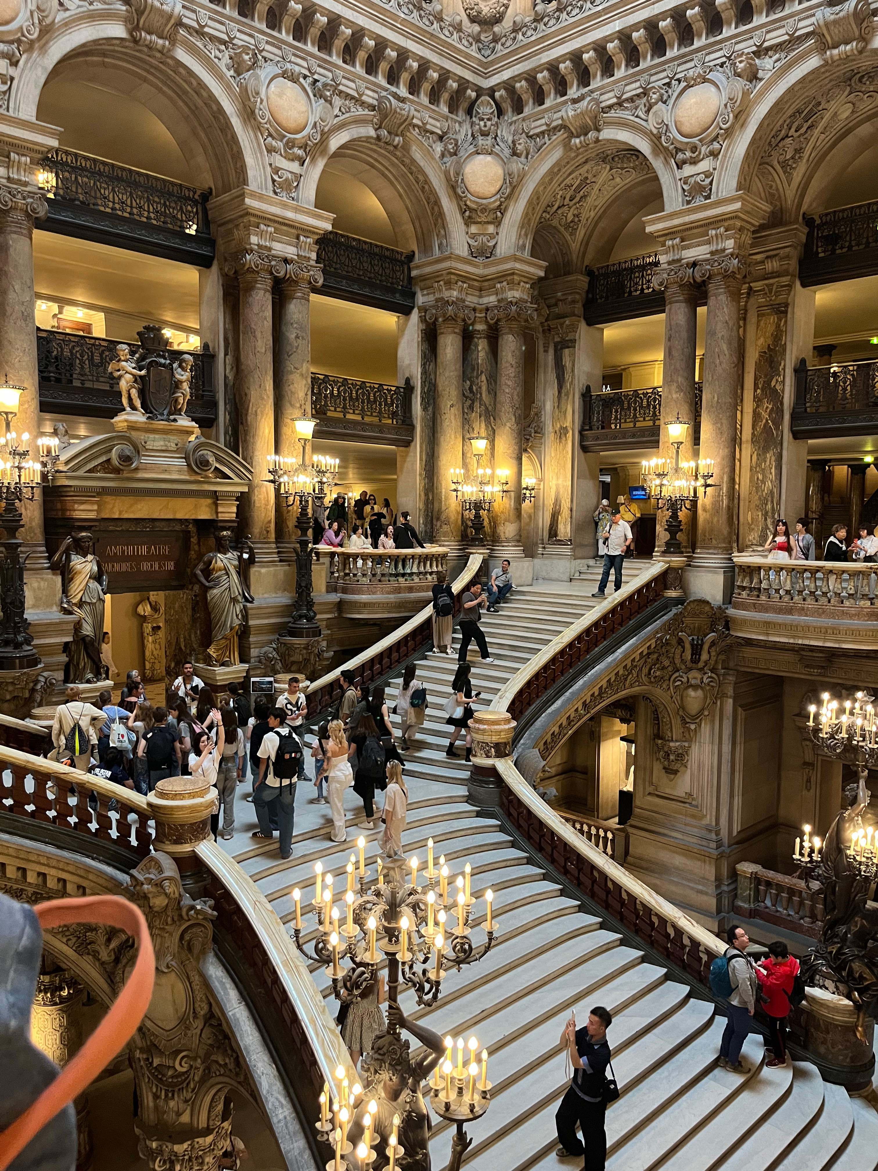 People walk up and down the grand marble staircase inside an ornate historic building with tall columns, arches, chandeliers, and detailed carvings.
