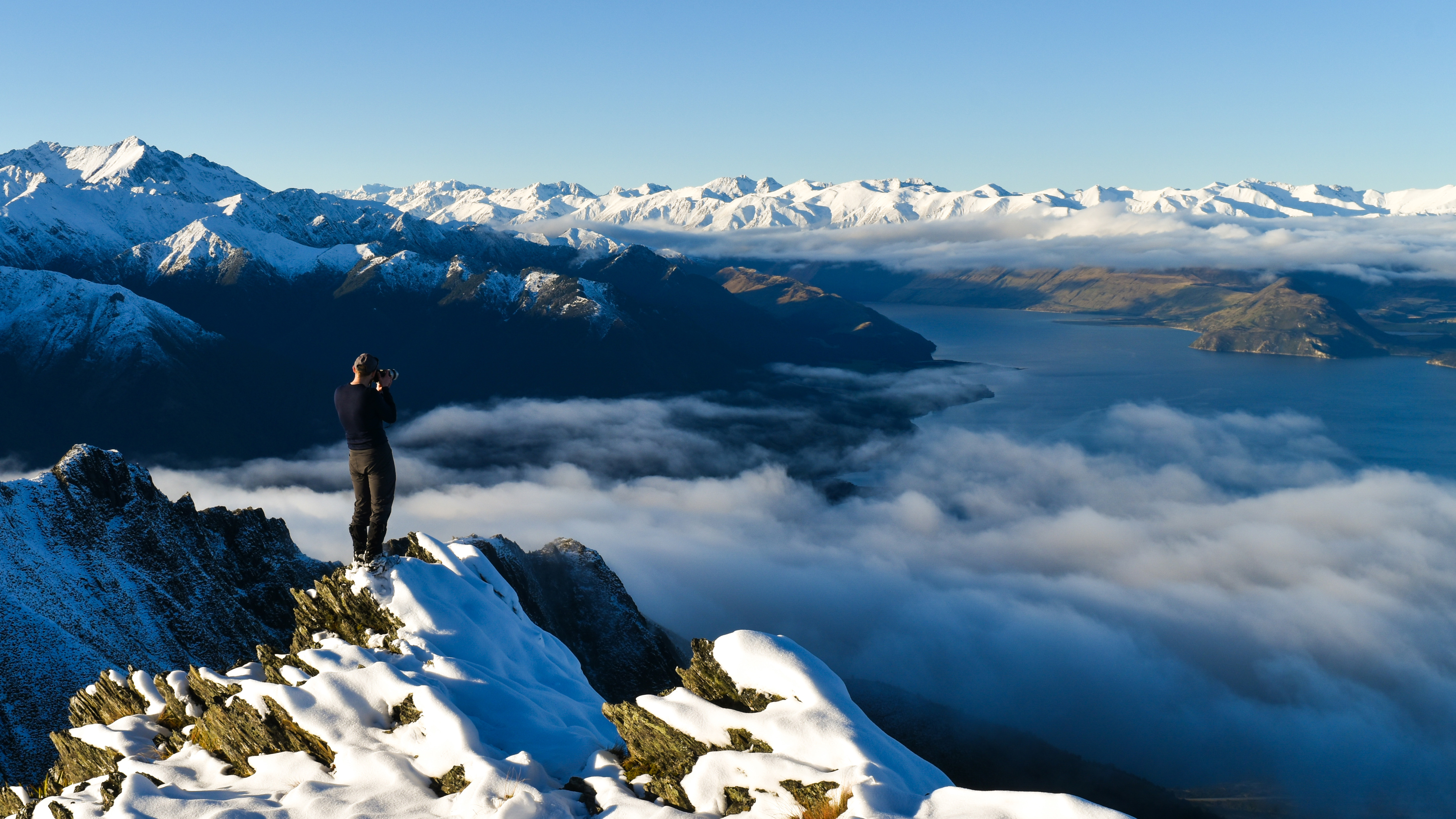 A mountaineer stands on a snow-covered mountain peak, taking a photo of a vast landscape of snowy mountains, clouds, and a lake below under a clear blue sky.