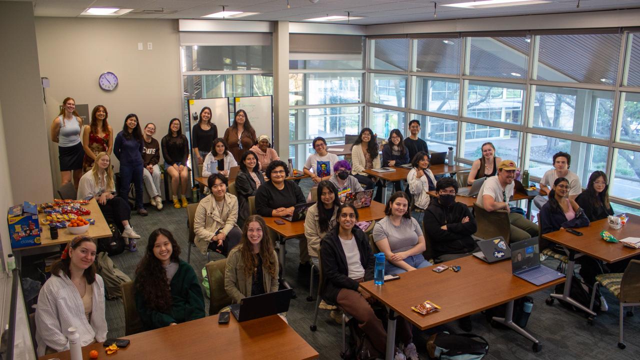A diverse group of students smiling in a classroom setting with tables and large windows.