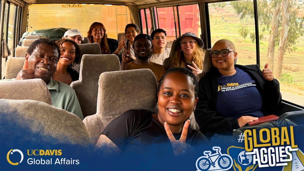 Group of smiling UC Davis students and partners posing on a bus, flashing peace signs and thumbs up.