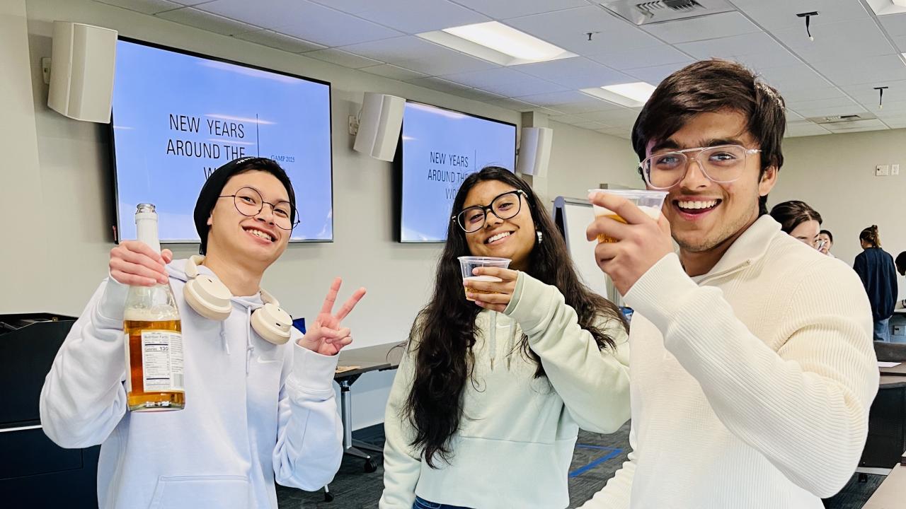 Three students smile and raise cups during a campus event, standing behind a table with fruit and snacks in a classroom setting.