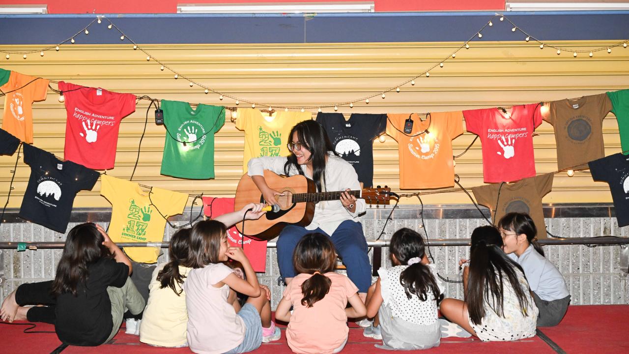 A person playing guitar and singing to a group of children seated on the floor in front of them. Colorful t-shirts and string lights hang behind them, creating a cheerful camp-like setting.