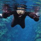 Two snorkelers in black drysuits swim in clear blue water above rocky underwater formations.