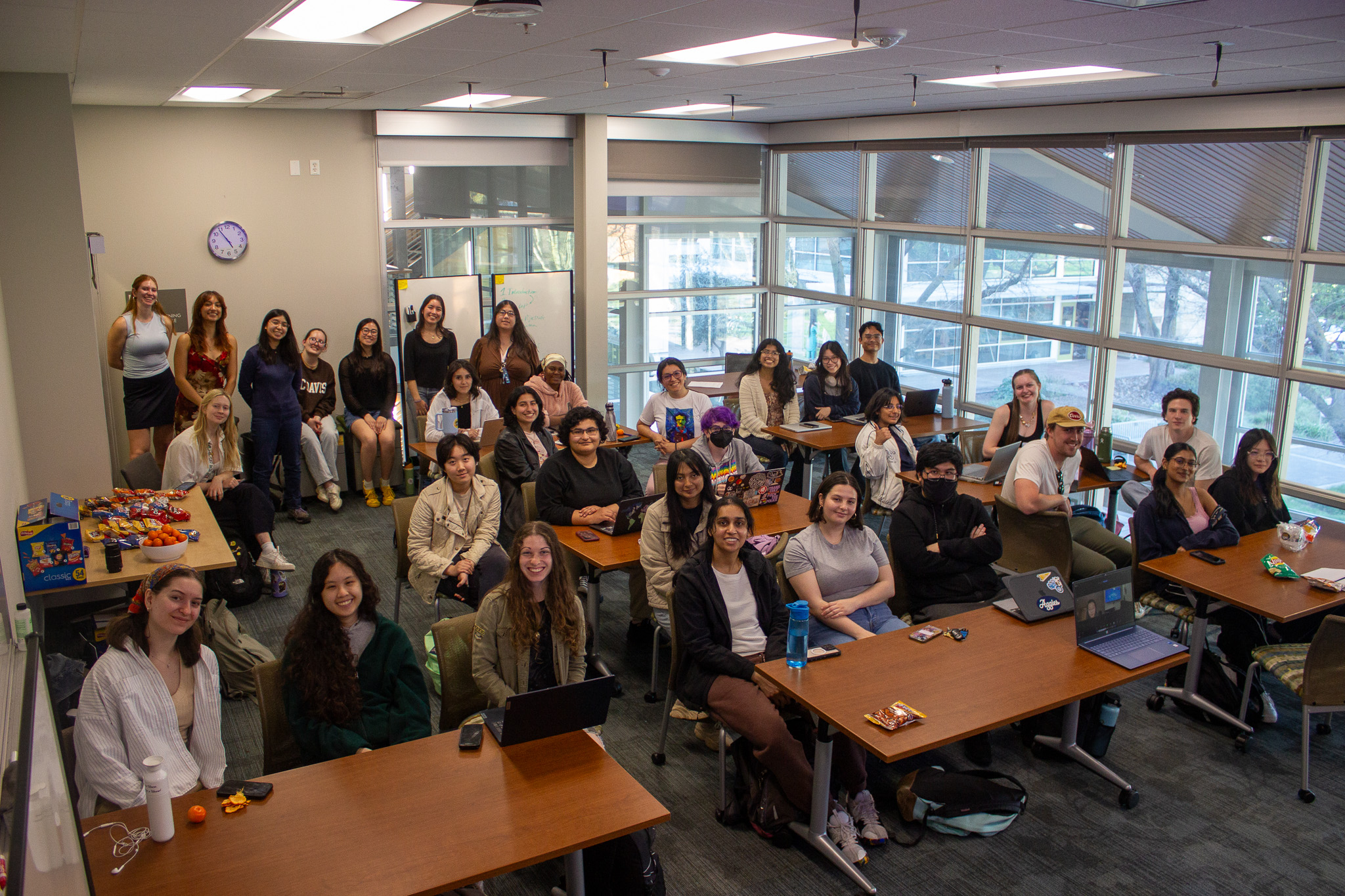 A diverse group of students smiling in a classroom setting with tables and large windows.