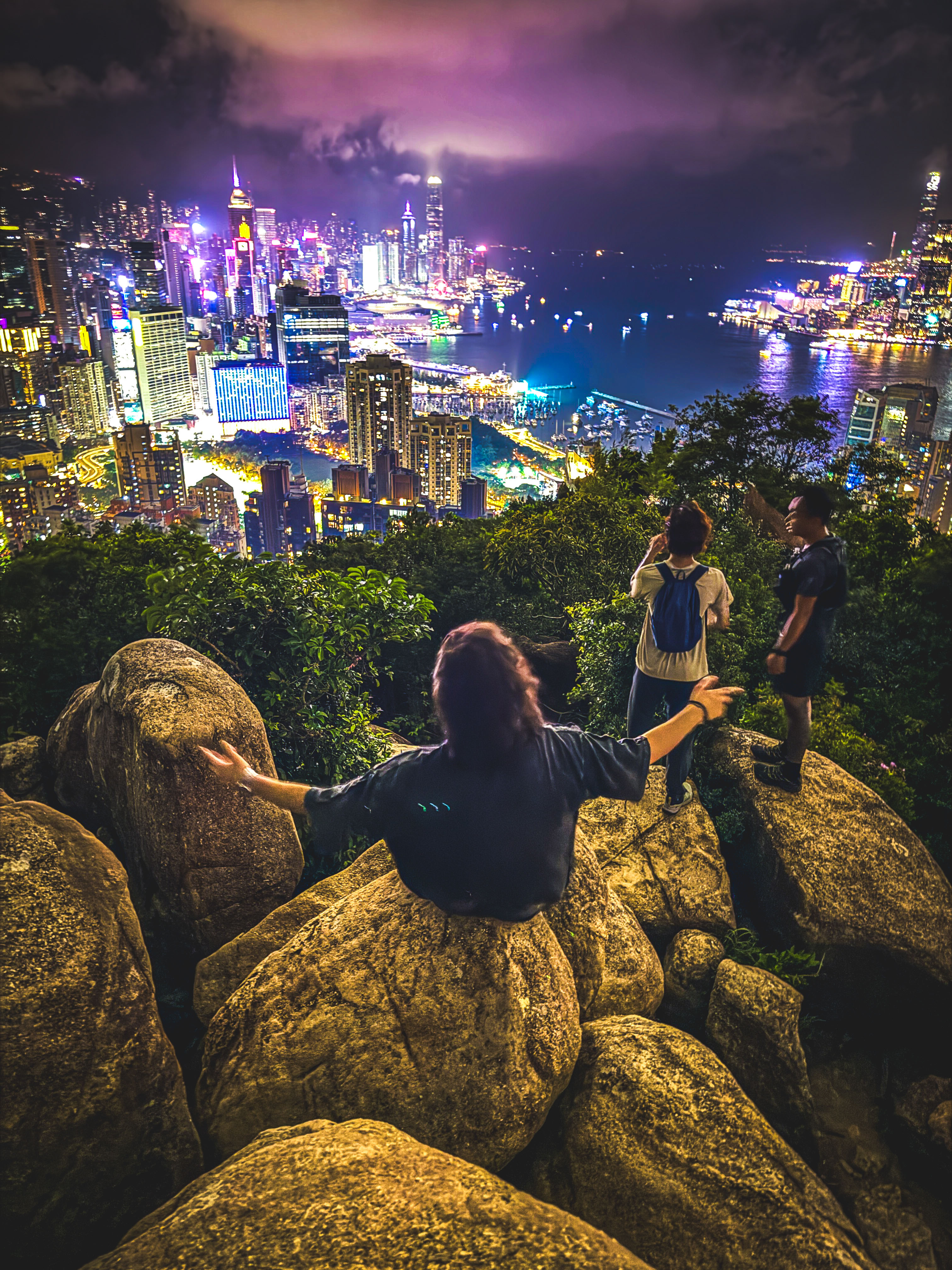 Three people stand on large rocks overlooking a brightly lit city skyline at night, with skyscrapers, water, and clouds glowing in the distance.