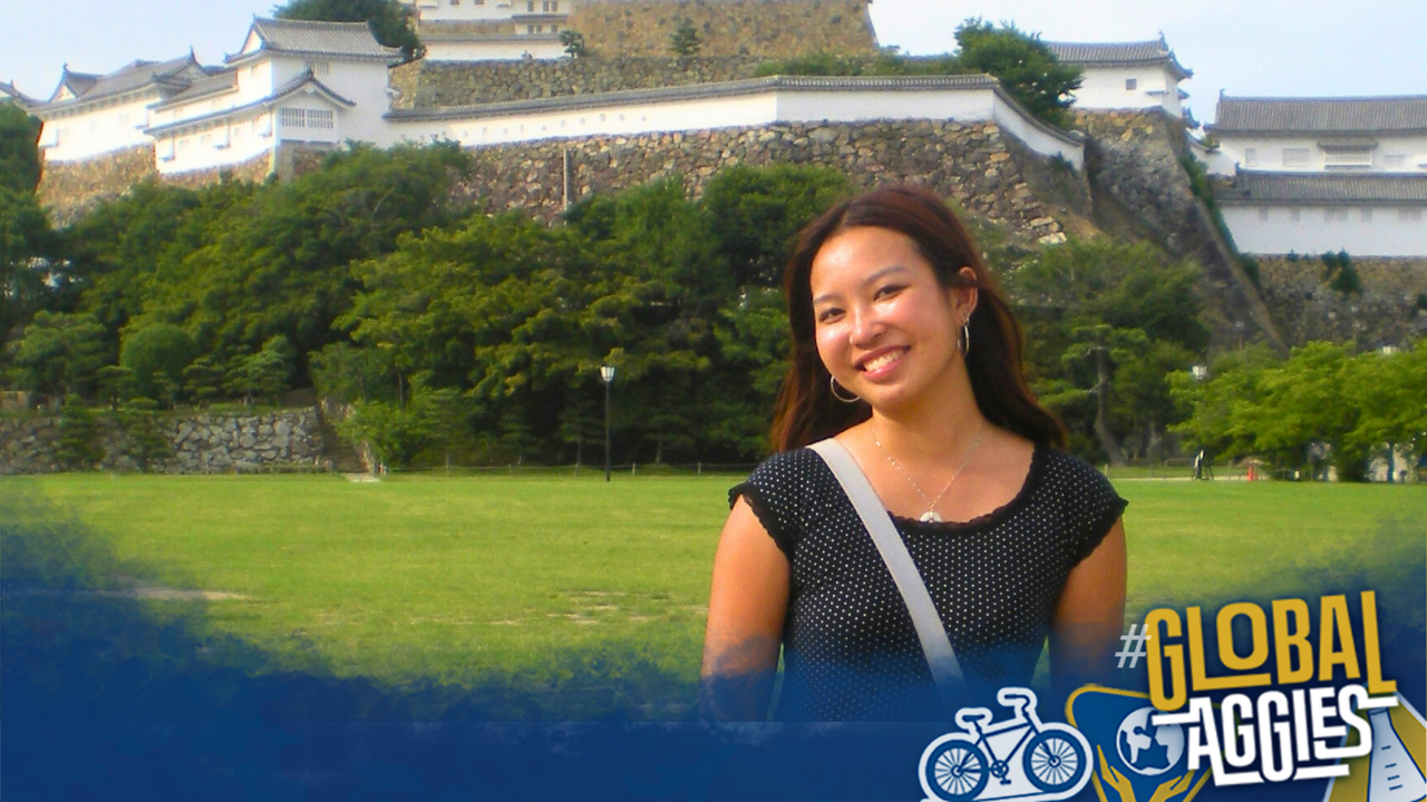 A smiling woman stands in a green field with a castle in the background.