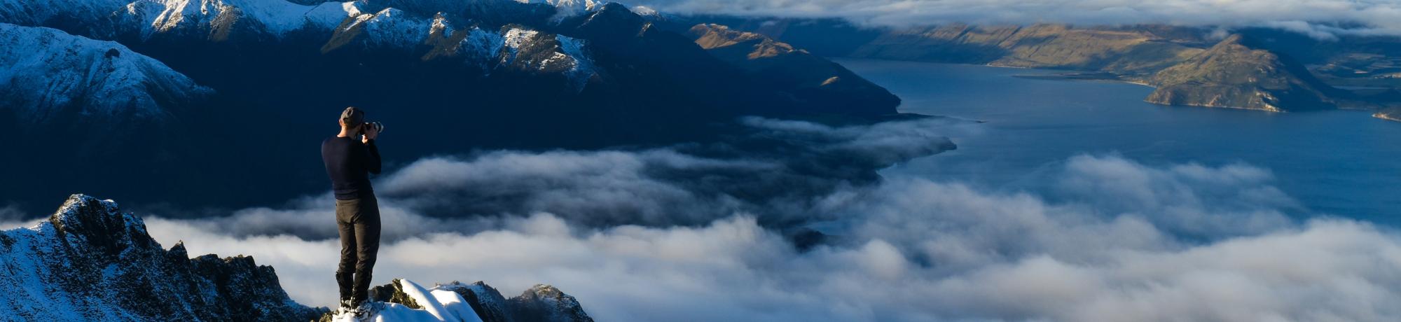 A mountaineer stands on a snow-covered mountain peak, taking a photo of a vast landscape of snowy mountains, clouds, and a lake below under a clear blue sky.